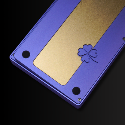 A detail photo of the What The Thock Lucky65 65% mechanical keyboard in the lavender color, showing the clover etching in the rear as well as the brass weight.