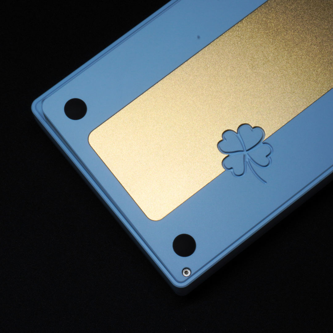 A detail photo of the What The Thock Lucky65 65% mechanical keyboard in the Ocean Blue color, showing the clover etching in the rear as well as the brass weight.
